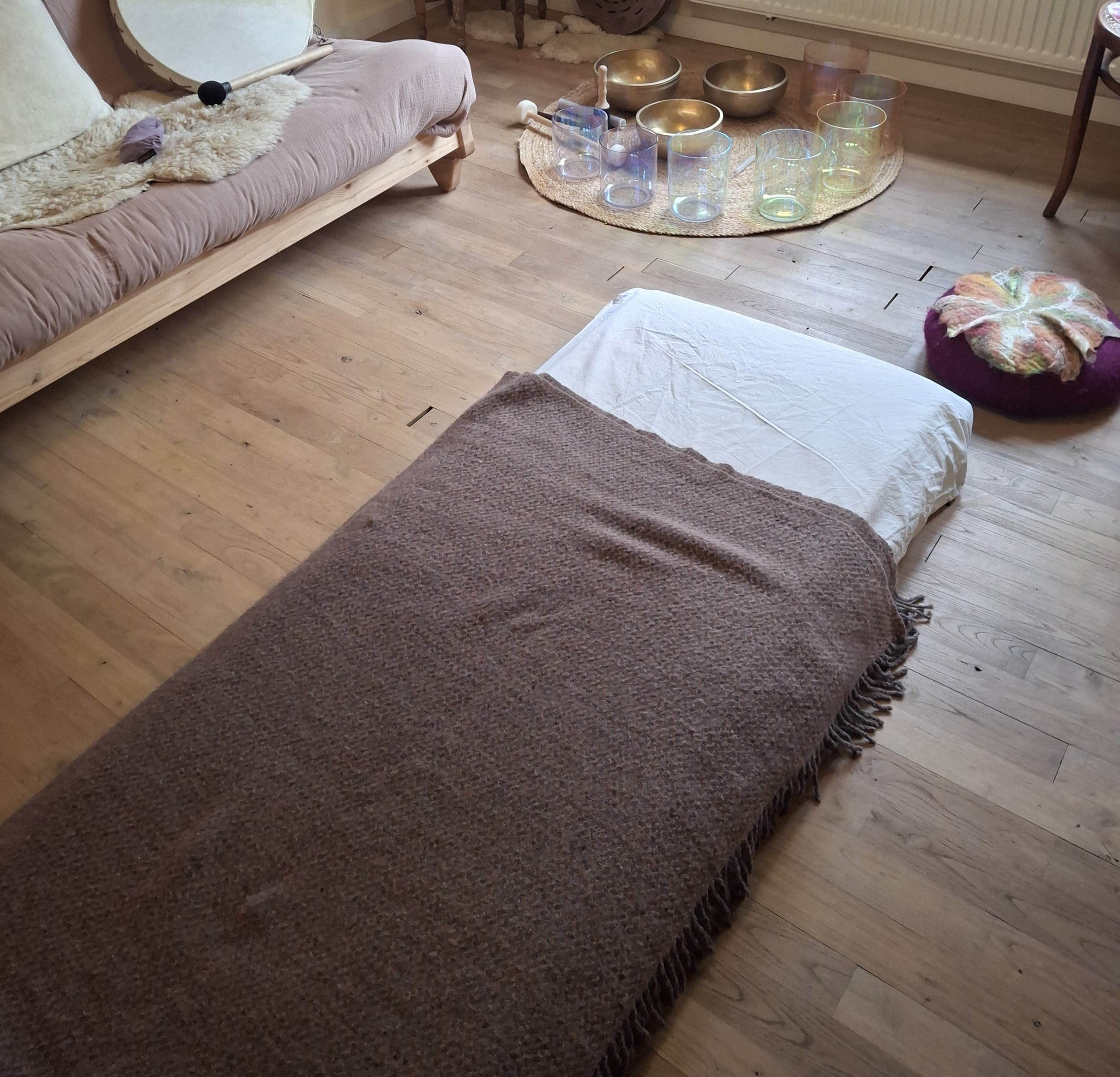 Een matras met een wollen deken op de vloer van de praktijkruimte, met klankschalen op de achtergrond